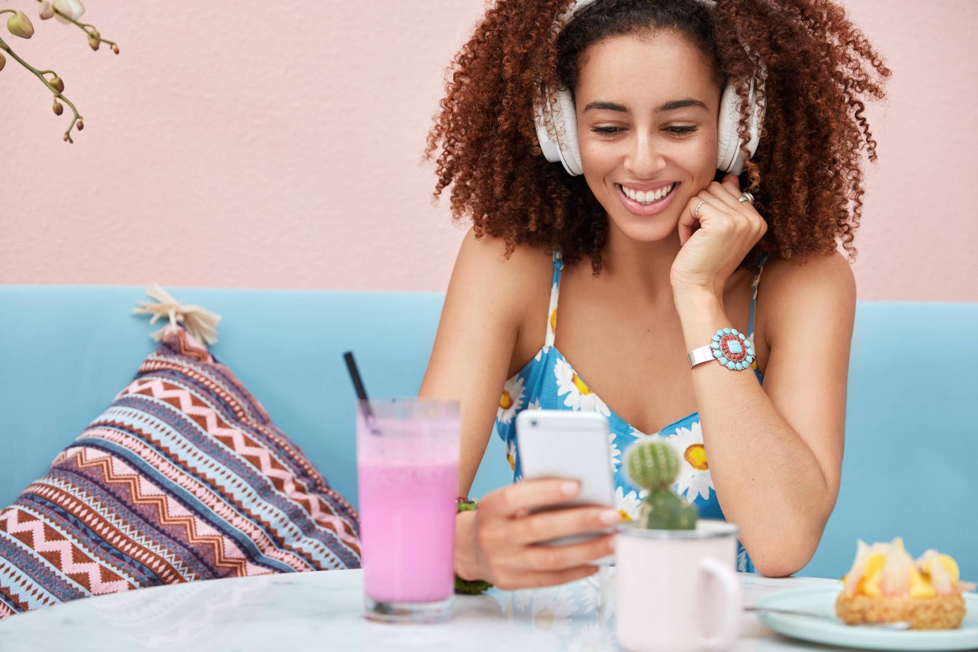 Horizontal shot of pleased African American dark skinned woman listens music from radio website, connected to wireless internet, drink smoothie and eats delicious cake, sits on sofa in coffee shop